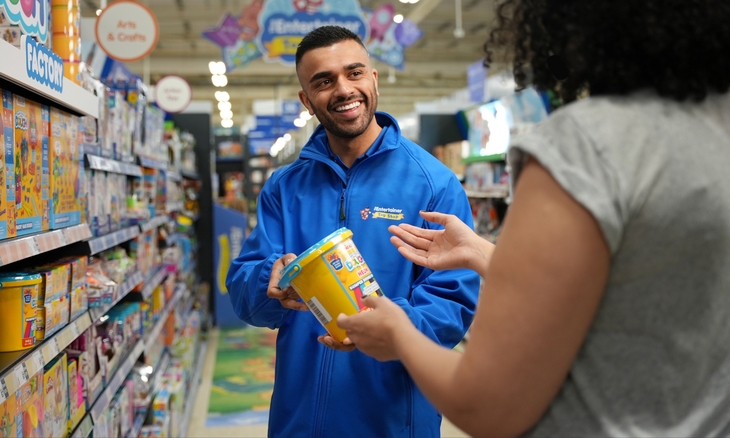 A smiling person of colour with short hair and stubble wearing a blue fleece helping a customer in a toy shop