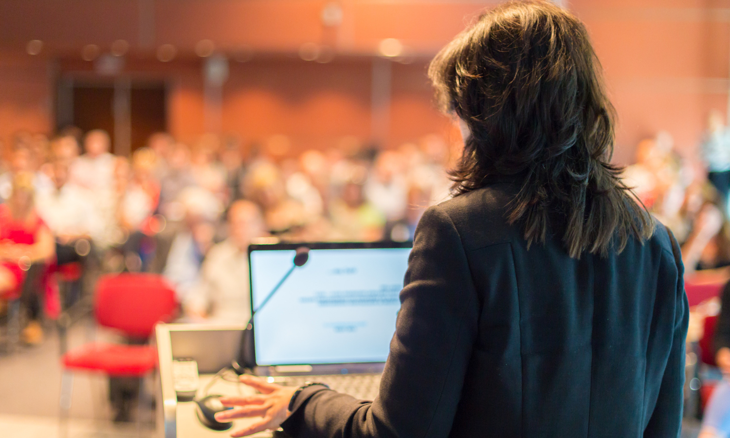 A person stands at a podium, delivering a lecture to a crowded room
